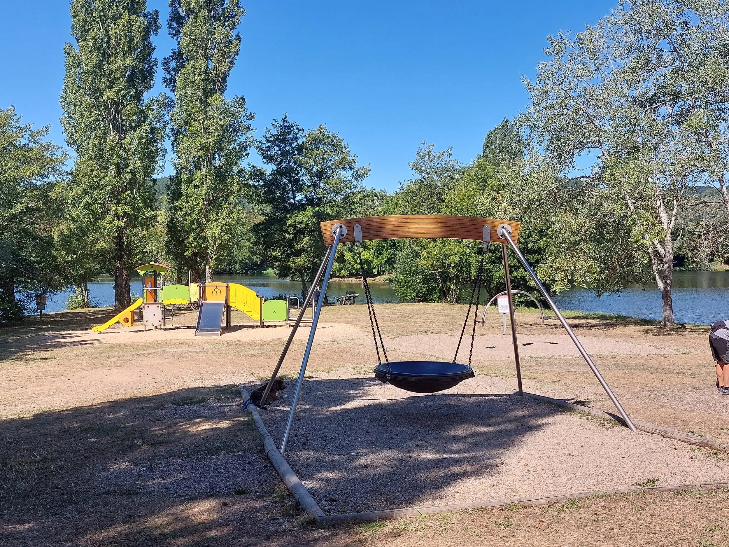 Children's playground with swing set in front of trees and a lake, with a playground structure featuring a slide and climbing area in the background.