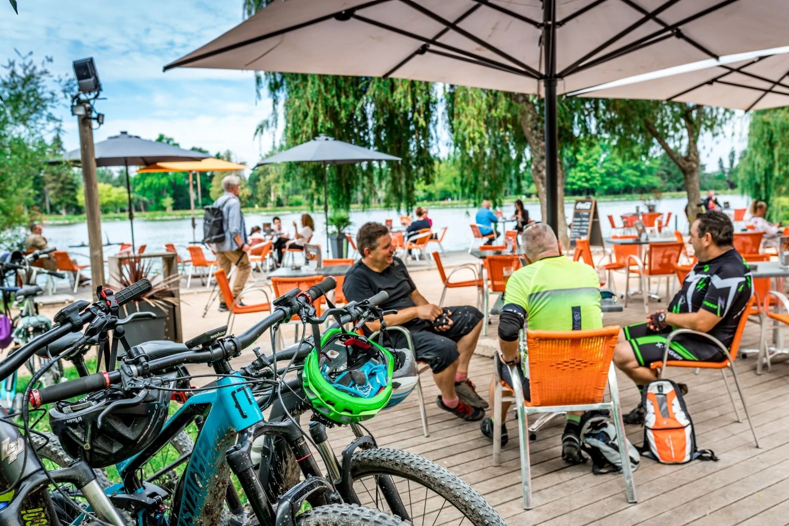Three cyclists resting at an outdoor cafe near a lake, with bikes in the foreground and people dining in the background.