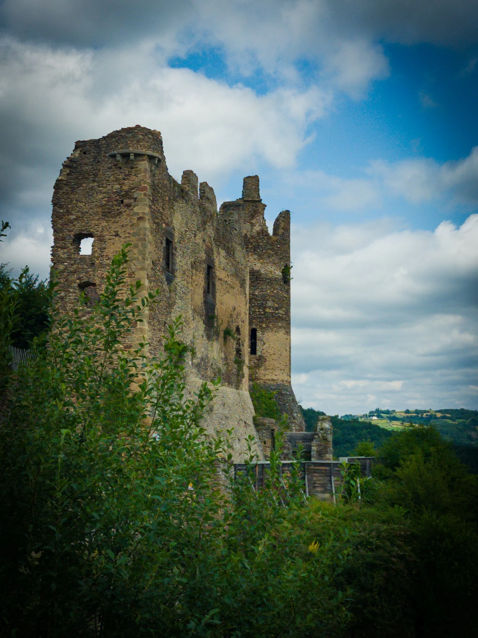 Ruined stone castle on a hill with green trees in the foreground and a cloudy sky in the background.