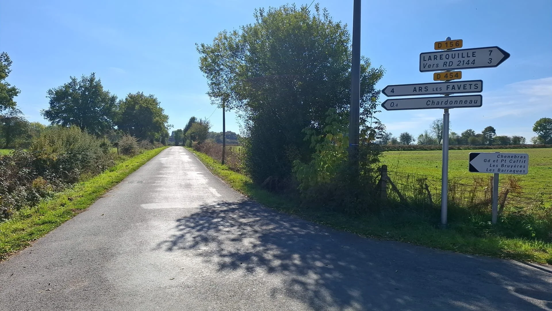 A rural road with multiple directional signs indicating nearby towns and routes. The road is flanked by green grass and trees, under a clear blue sky.