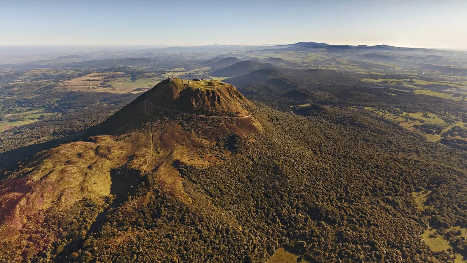 Aerial view of a mountain surrounded by green fields and forests, with a small structure and a wind turbine at the summit.
