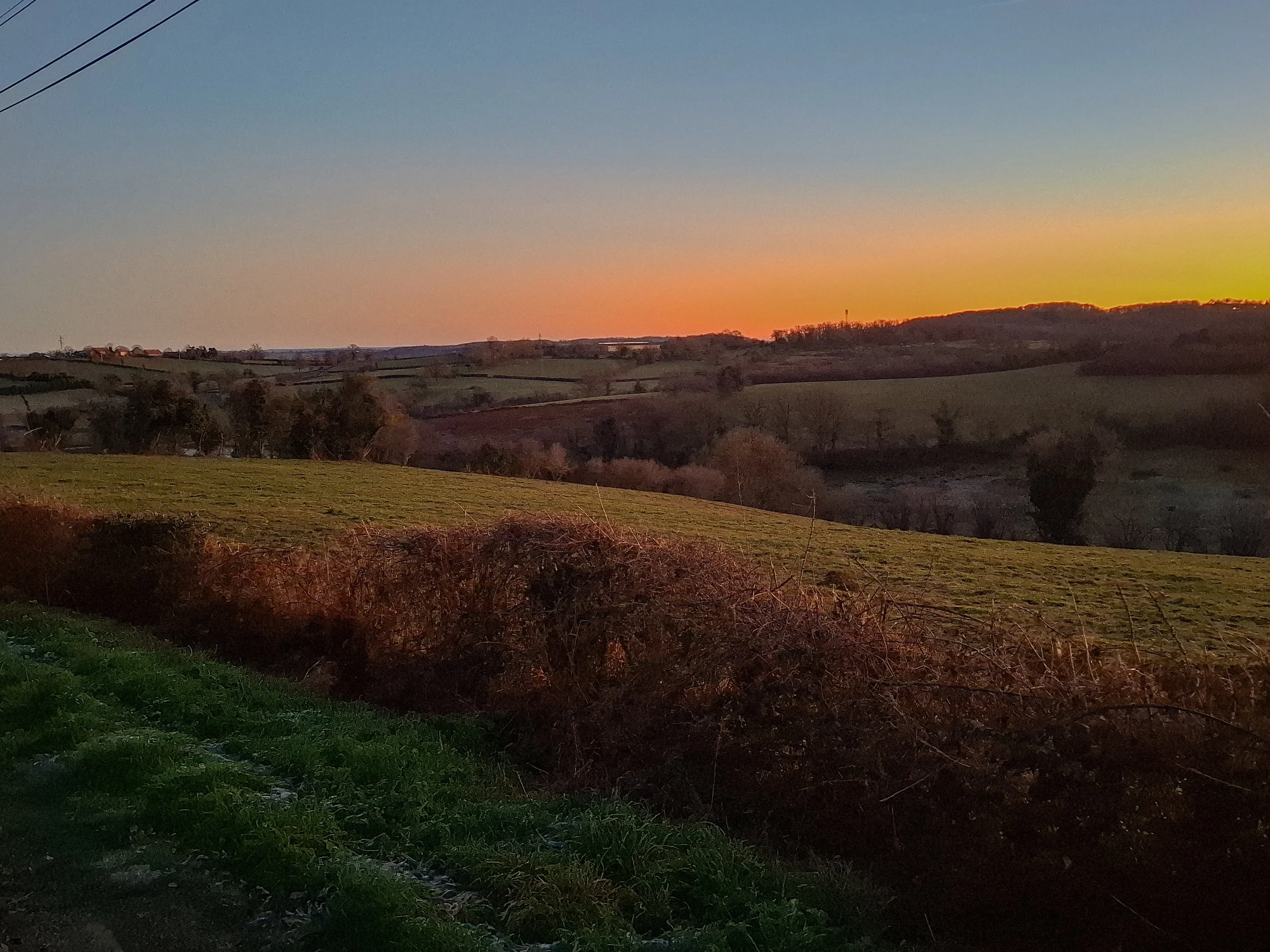 A peaceful countryside landscape at sunset with rolling hills, trees, and a colorful sky.