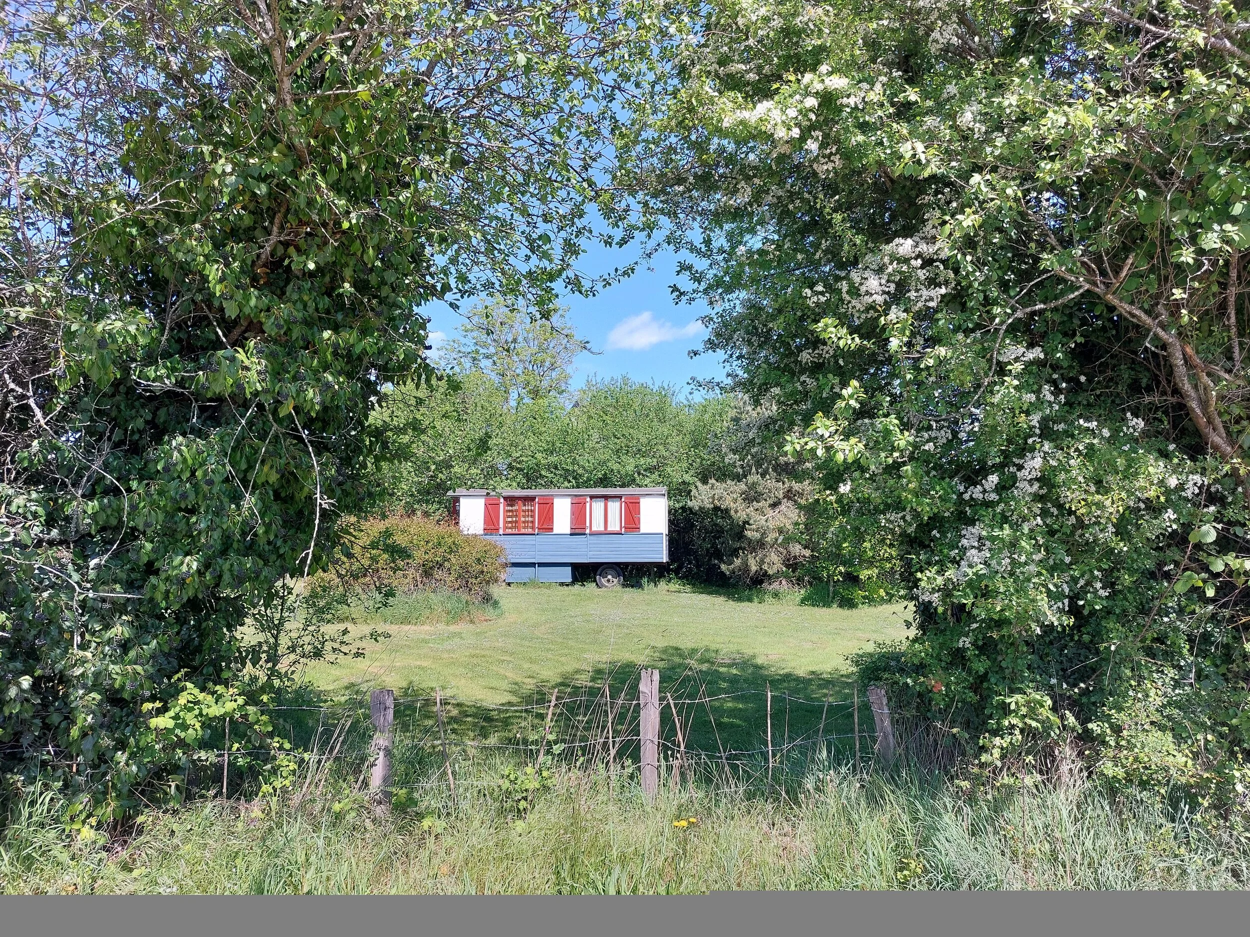 A mobile home with white walls and red shutters is situated among green trees and bushes, viewed through a grassy area with a wooden fence in the foreground on a sunny day.