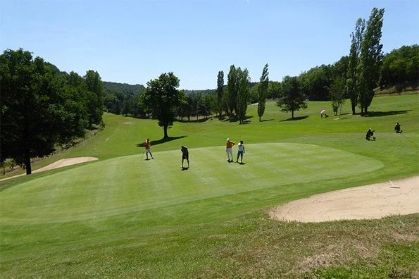 Golf course with players practicing near a sand trap, surrounded by trees and hills under a clear sky.