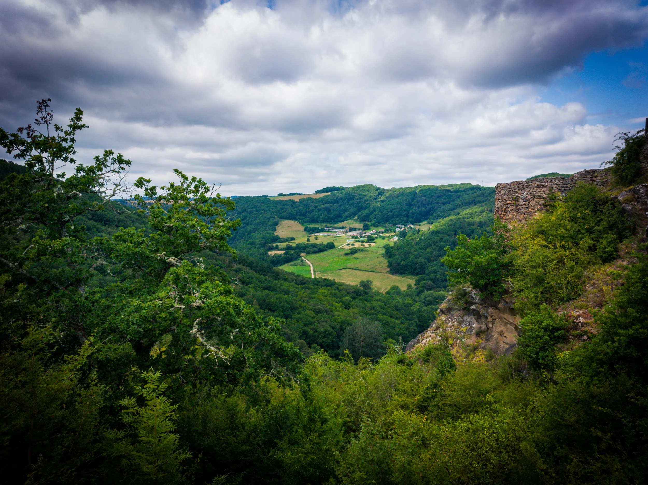 A lush, green valley surrounded by rolling hills under a cloudy sky, with a stone structure visible on the right side.