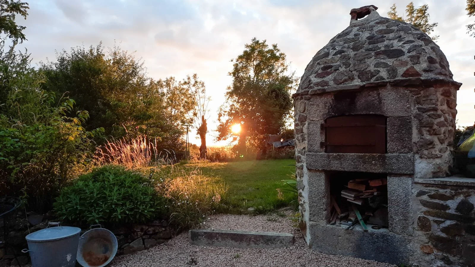 A stone outdoor oven or fireplace in a garden during sunset, with trees, bushes, and a grassy lawn in the background, and gardening tools and buckets in the foreground.