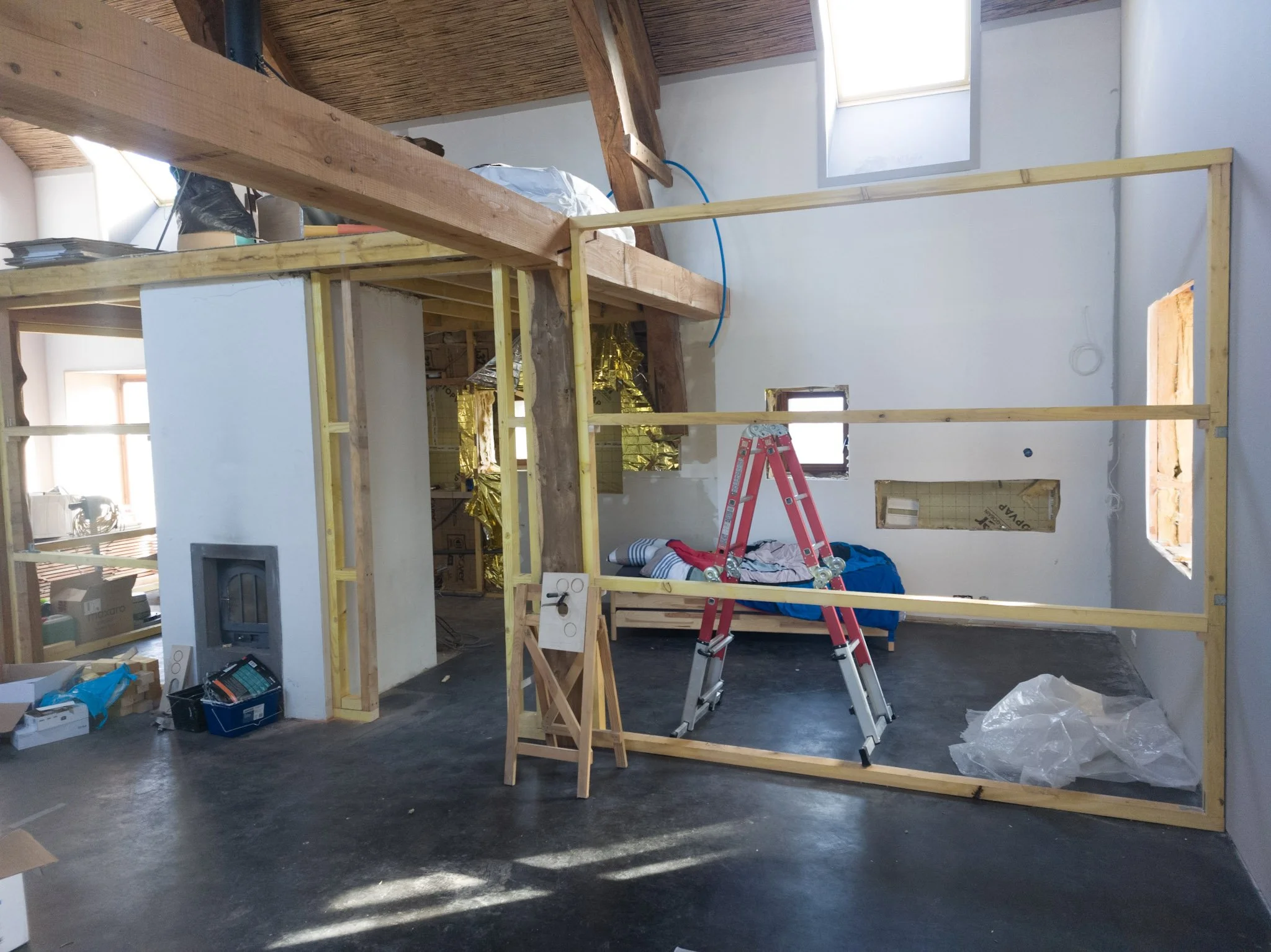 The interior of a room under construction with wooden framing, a red ladder, construction tools, and materials visible. Finished walls and windows are also present.