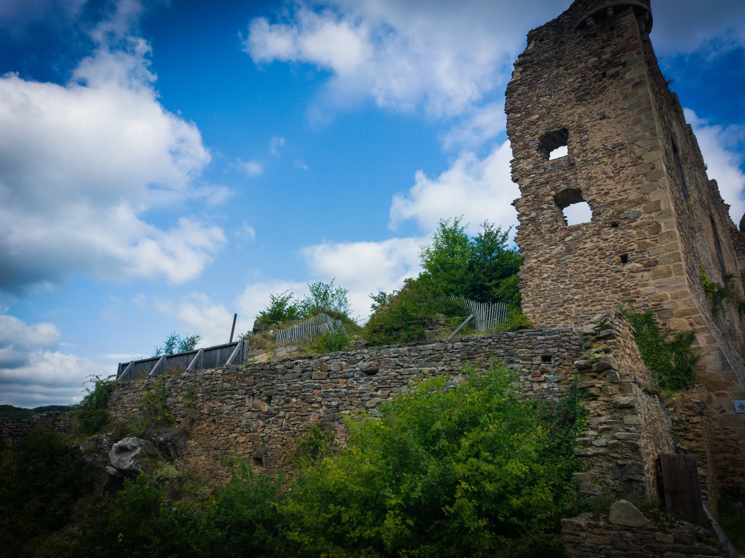 Ruins of an old stone building on a hill with a wooden fence and green trees, under a partly cloudy blue sky.