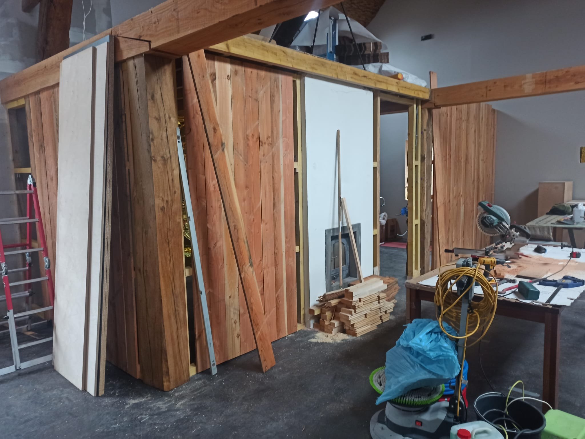 Construction site with wooden framing and tools, including a circular saw, ladder, and various pieces of wood, indicating ongoing building work.
