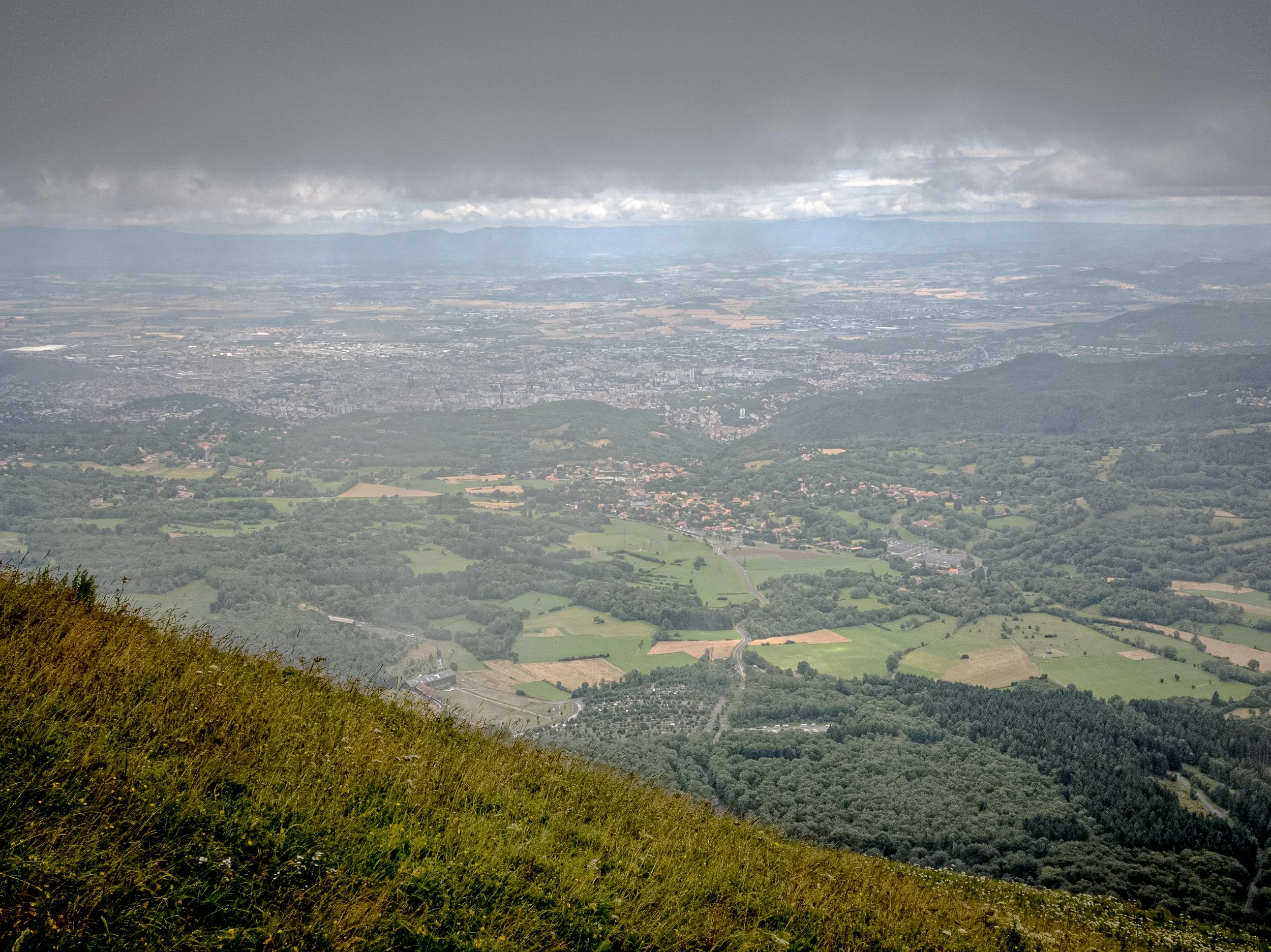 A view from a mountain showing a green landscape with fields, forests, and small roads, with an urban area in the distance under a cloudy sky.