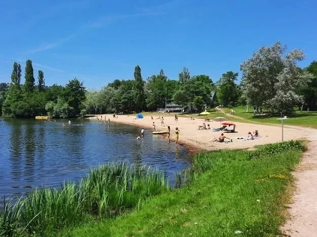 People relaxing and swimming at a sandy beach by a lake on a sunny day, with trees and houses in the background.