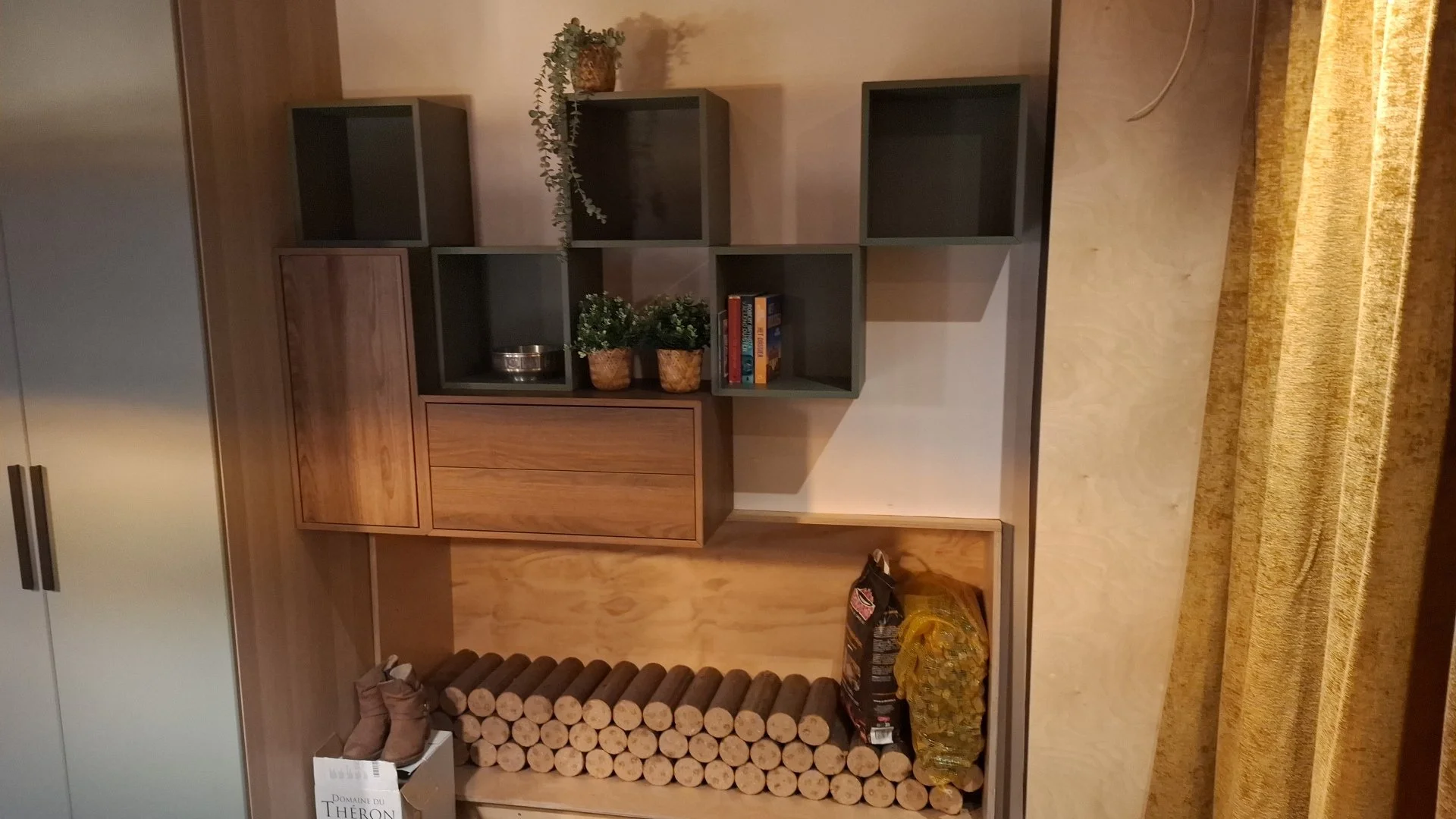 A modern room corner with a wooden and gray shelving unit, decorated with plants and books. Below is a stack of firewood, a cardboard box with shoes, and a large bag of chips. Part of a gold-colored curtain is visible on the right.