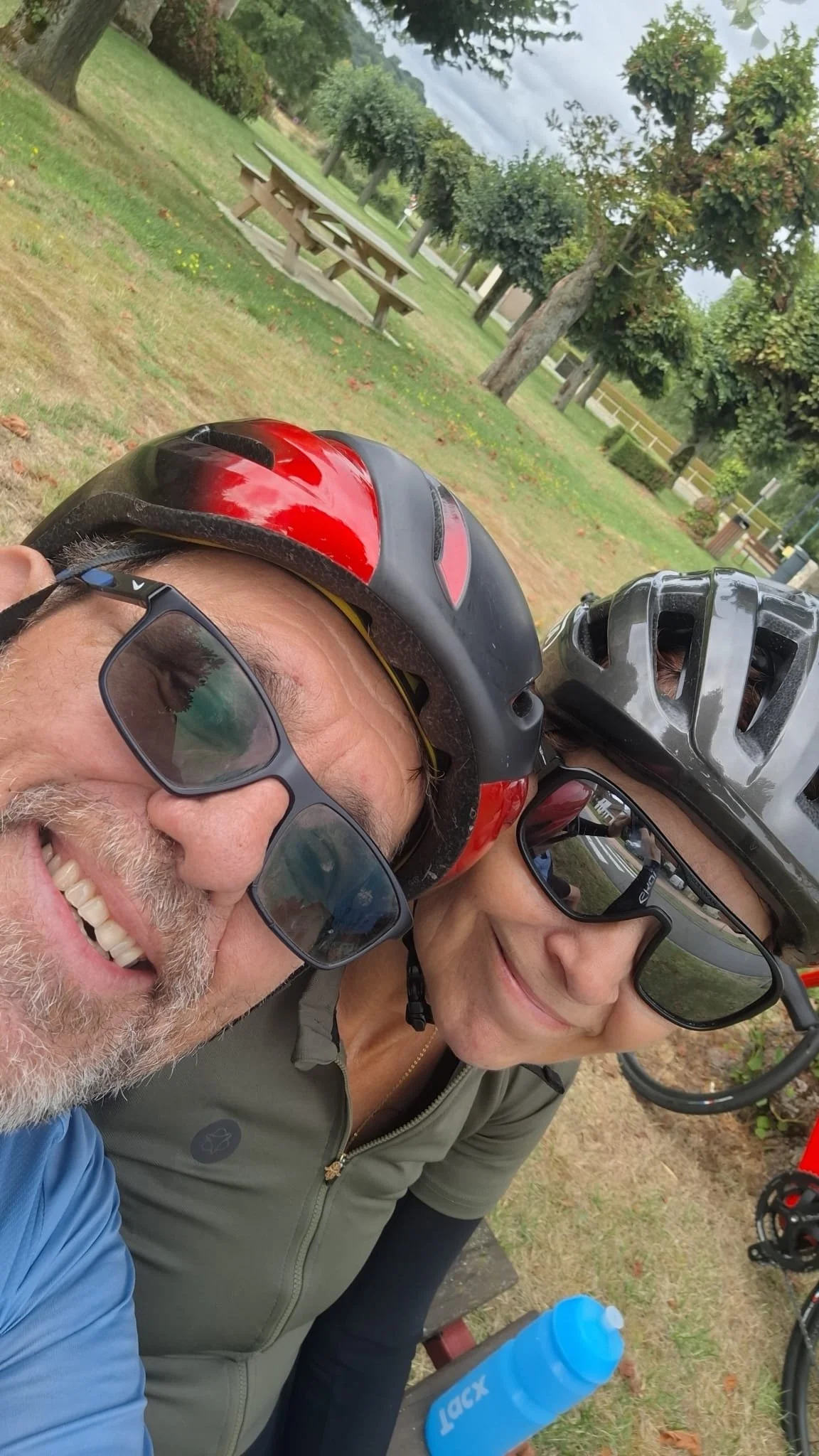 Close-up of a smiling man and woman wearing helmets and sunglasses, taking a selfie outdoors in a park with trees and picnic tables.