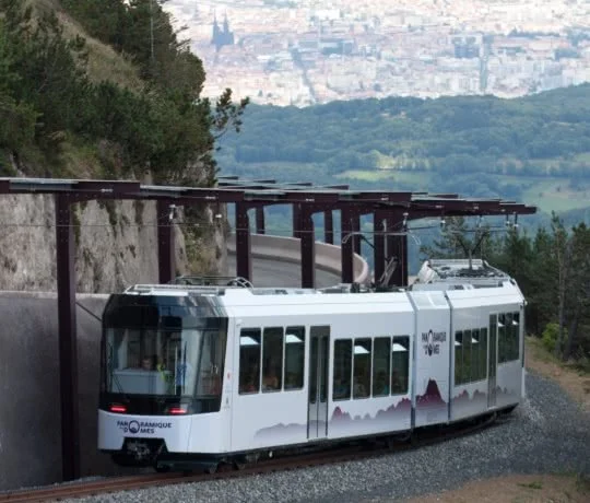 A white tram on a mountain track with a forest and cityscape in the background.