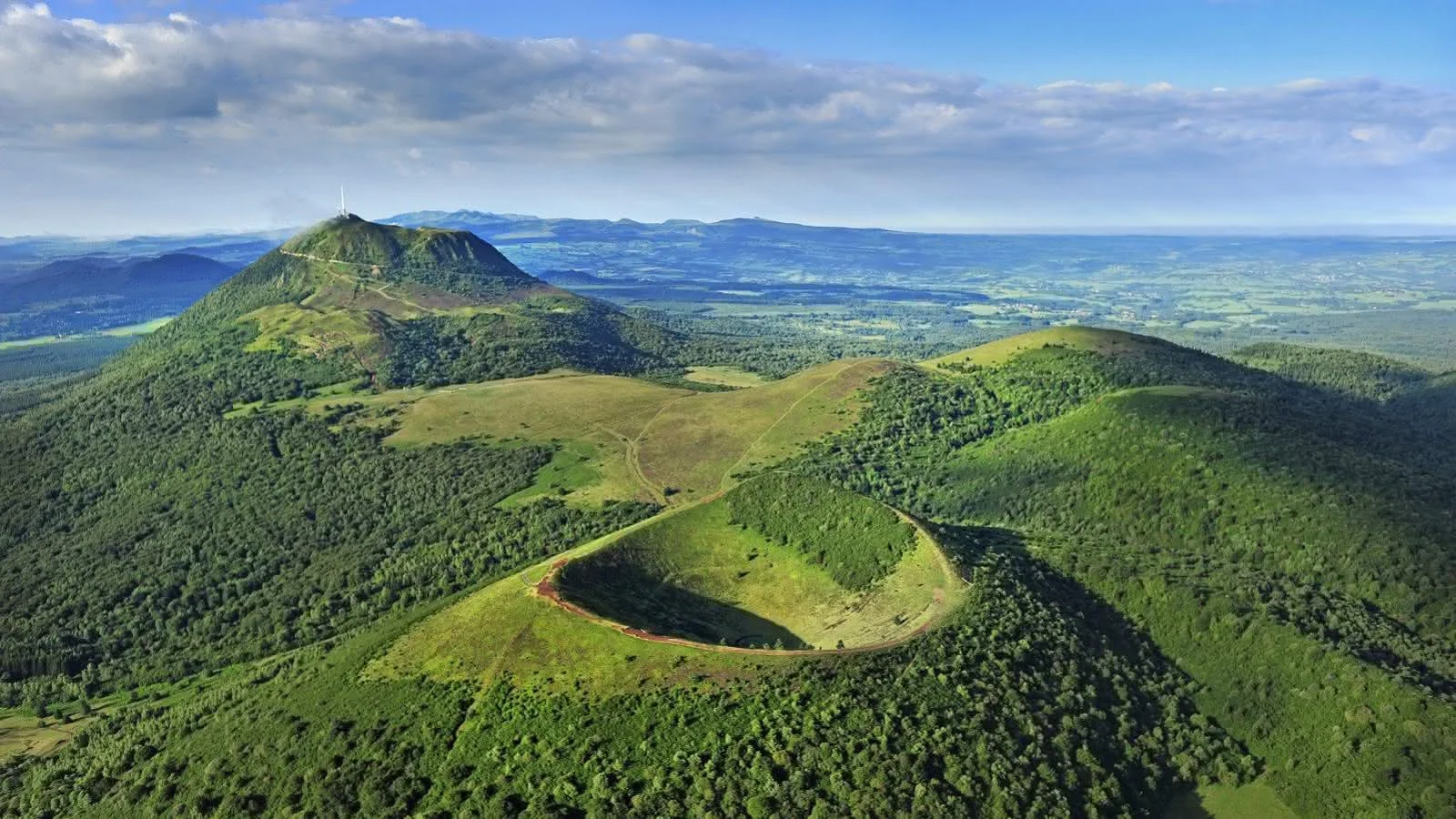 Aerial view of a lush green mountain landscape with winding trails and a hilltop tower under a partly cloudy sky.