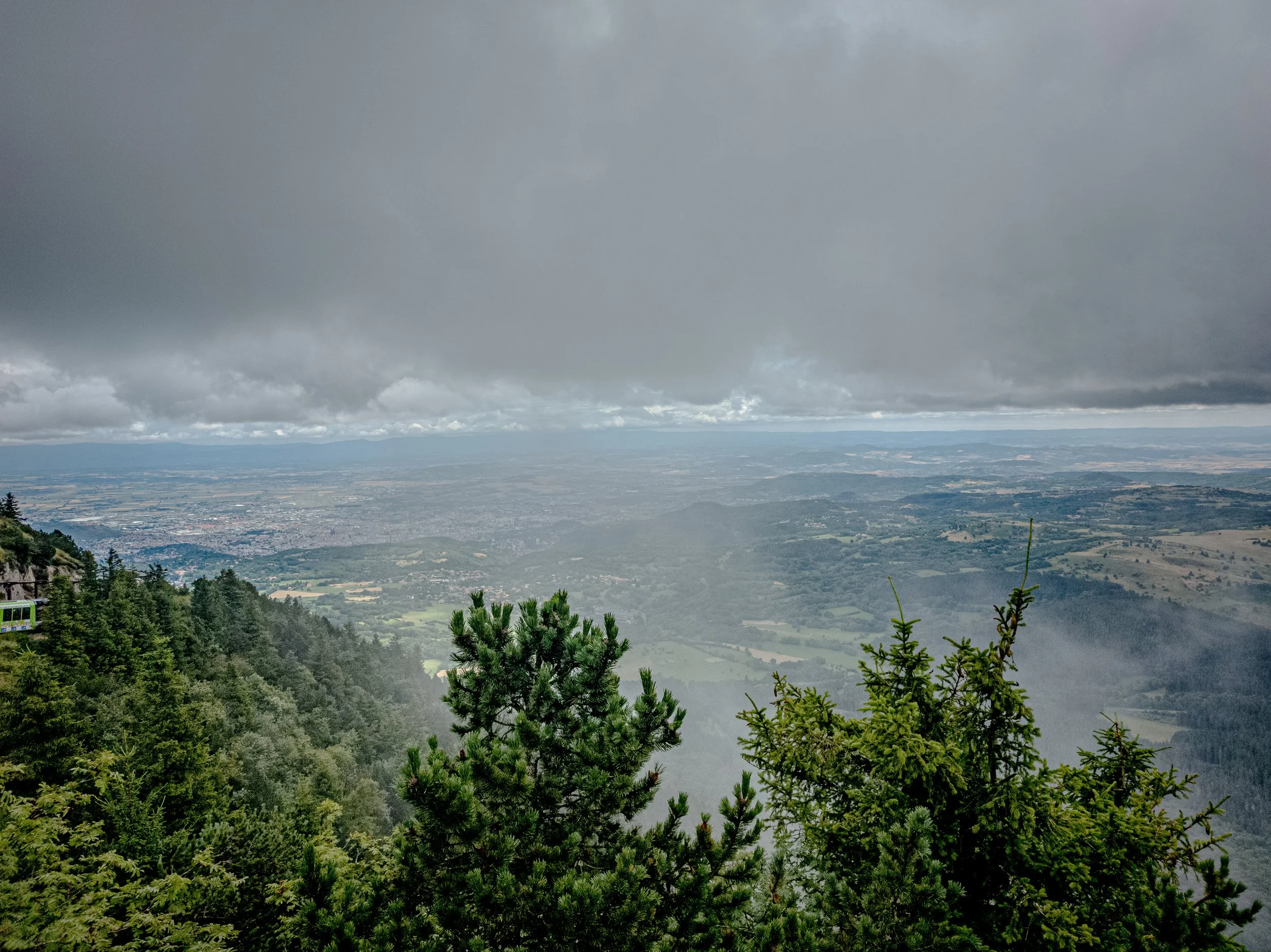 View of a mountain landscape with trees in the foreground, mist, and a city in the distance under a cloudy sky.