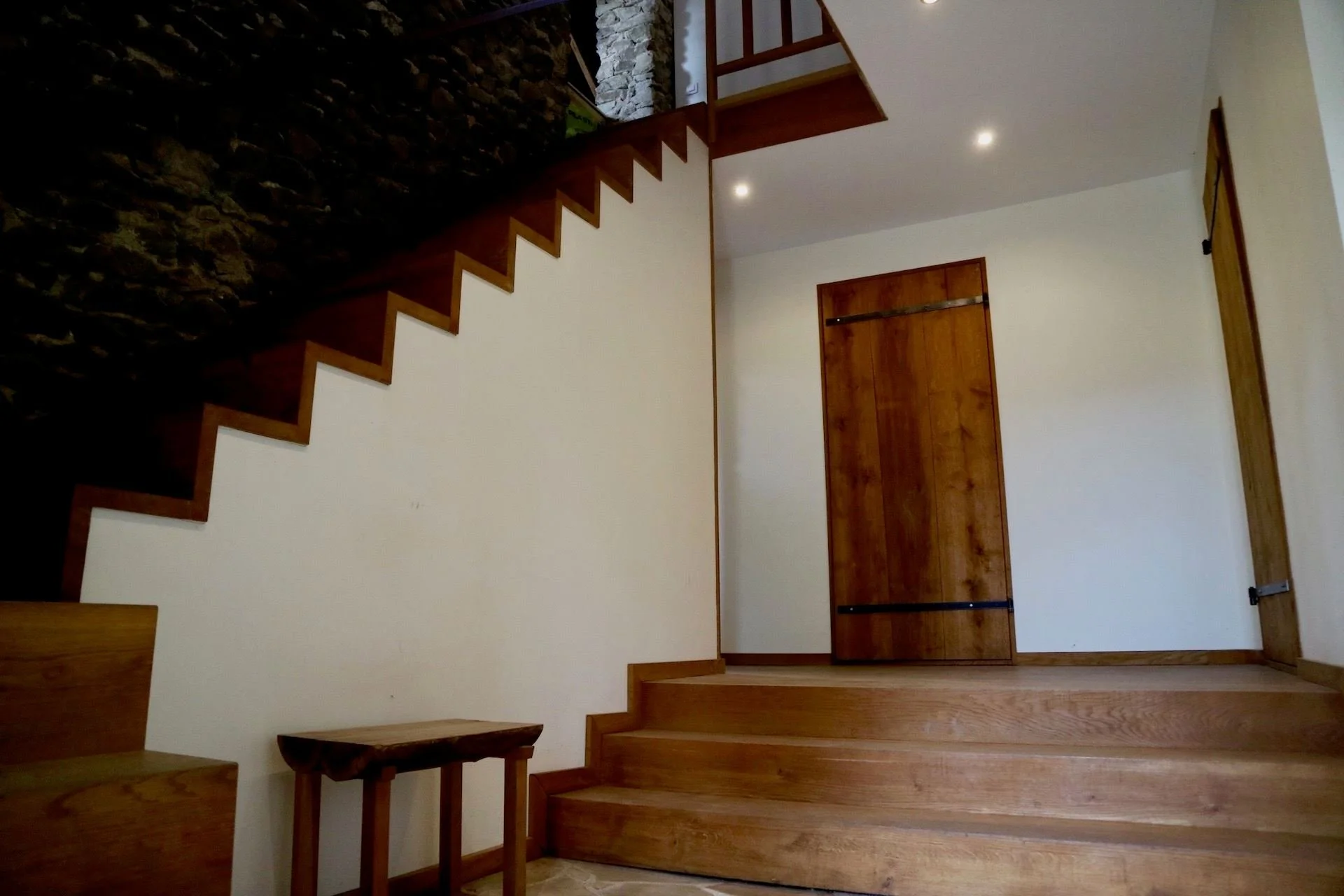 Interior view of a staircase with wooden steps and a door on the wall, with ceiling lights and a small wooden bench.