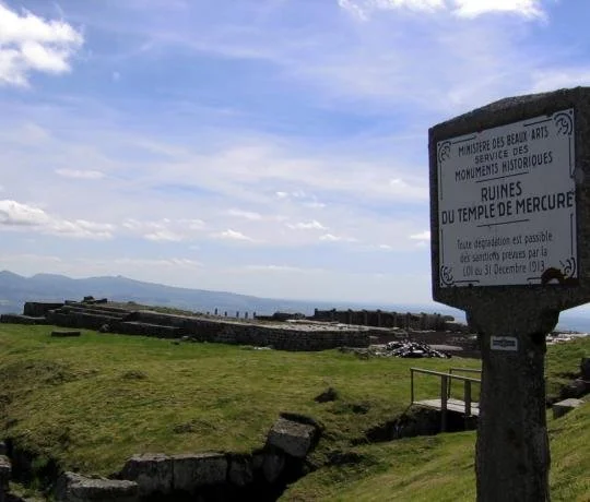 Ancient ruins of the Temple of Mercury on a grassy hill under a partly cloudy sky, with a sign in French describing the site.