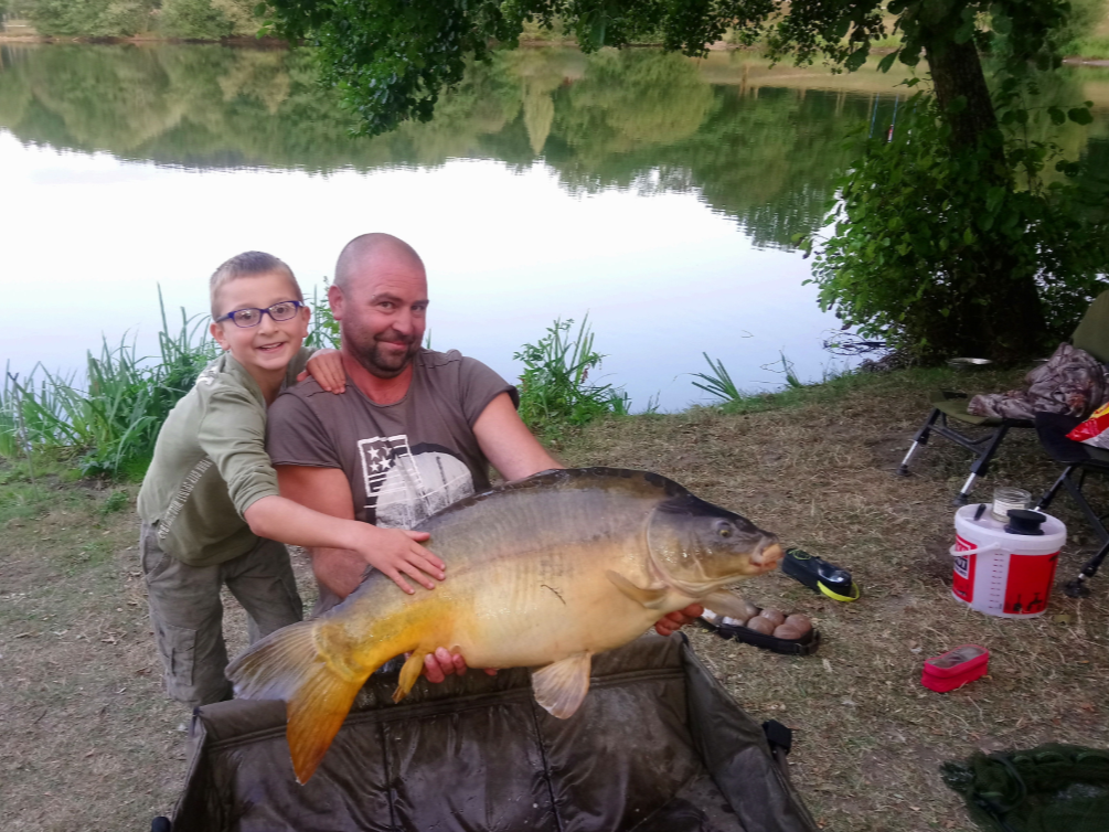 A man and boy holding a large fish by a lake, with fishing gear and trees in the background.