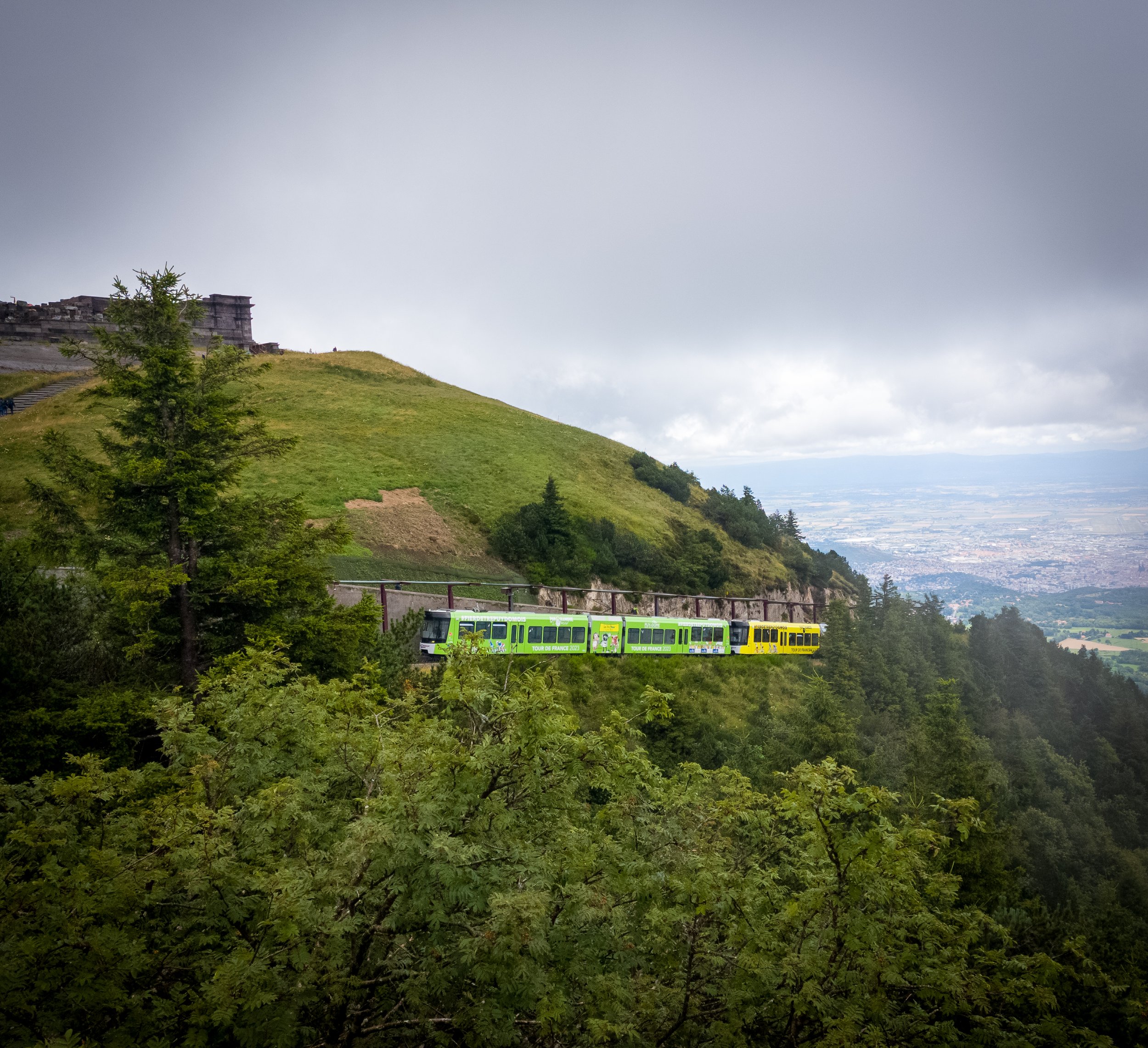 A green and yellow cable car traveling along a track on a mountainside, surrounded by green trees and hillside, with a cloudy sky overhead.