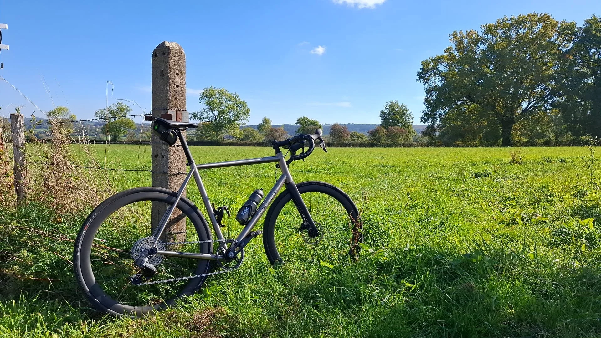 A bicycle leaning against a fence post in a grassy field on a sunny day with clear blue sky and trees in the background.
