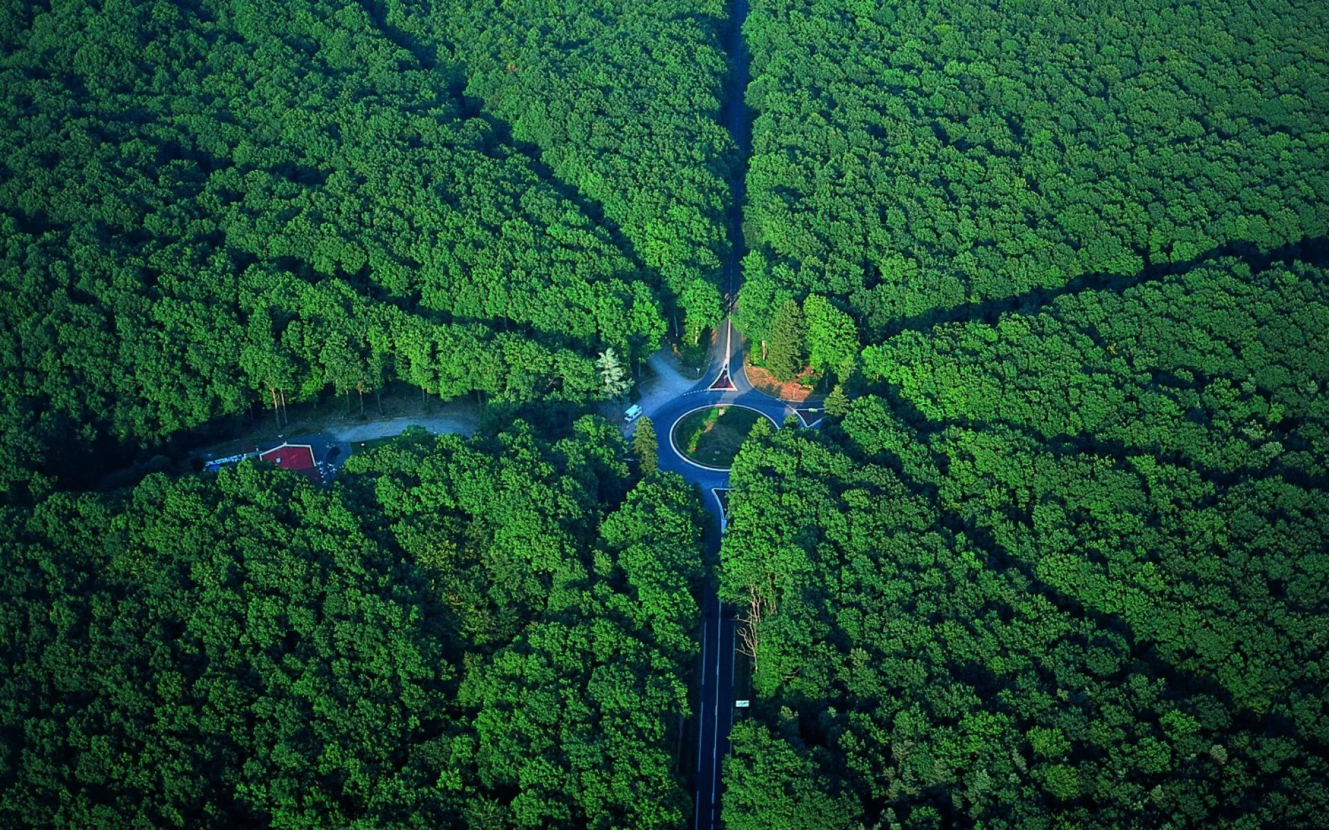 An aerial view of a roundabout highway intersection surrounded by dense green forest with a small building visible near the left side of the image.