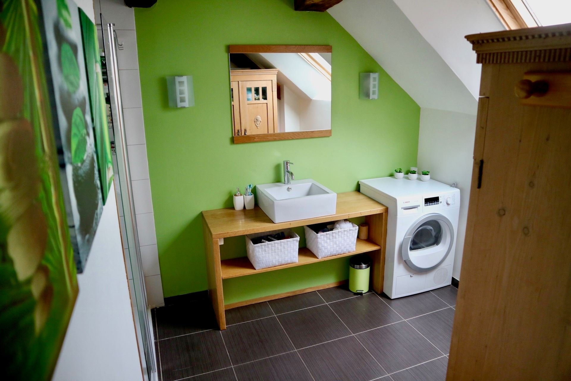 Laundry room with lime green wall, white washing machine, wooden countertop with a white sink, mirror above, small potted plants on the right, and brown tiled floor.