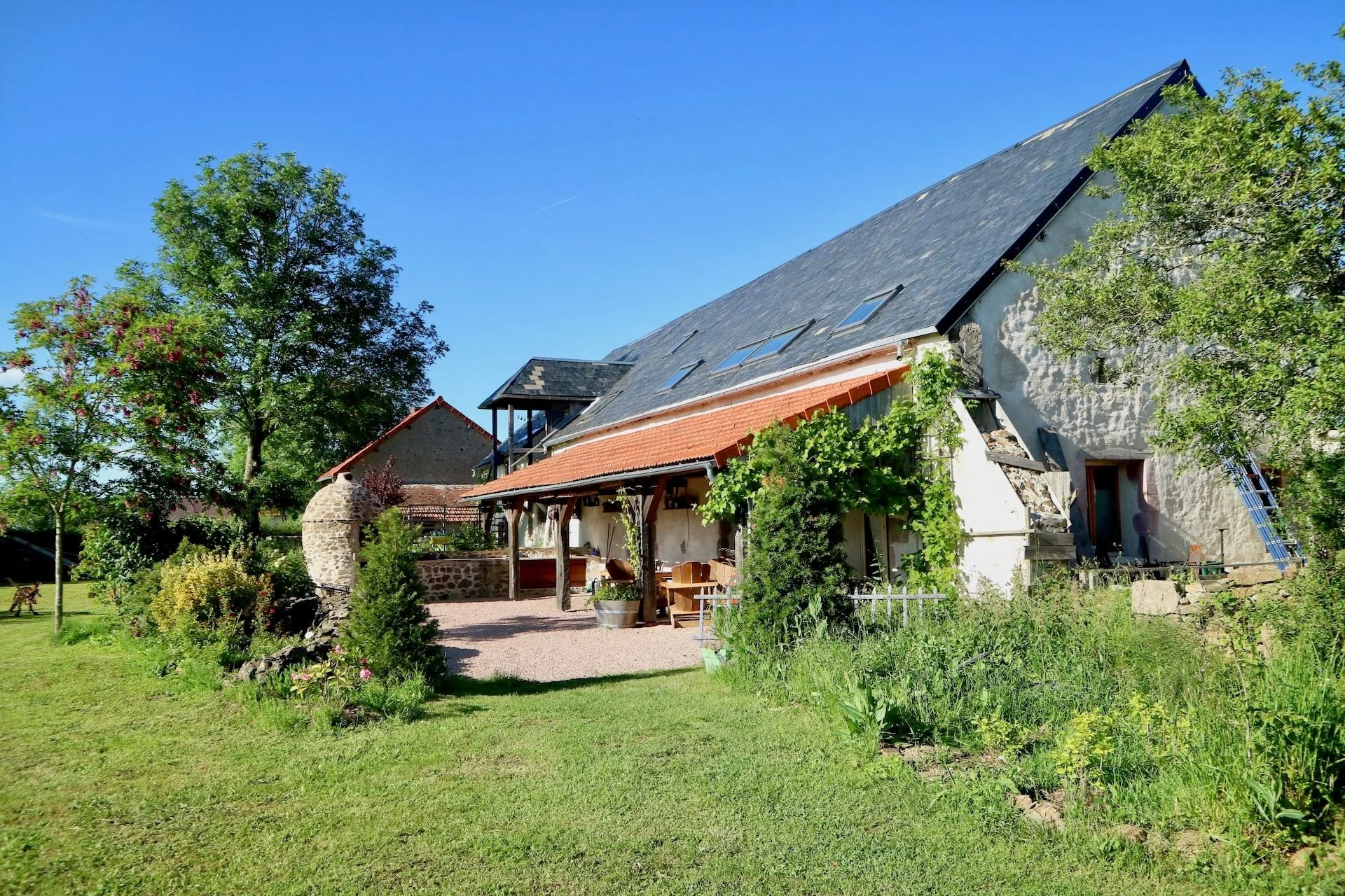 A rustic house with a sloped roof, outdoor patio area, surrounded by a lush green yard with trees and plants, under a clear blue sky.