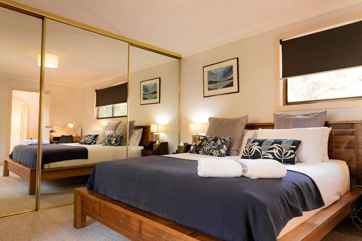 A cozy bedroom with two beds, large mirrored closet doors, wall art, nightstands with lamps, and windows with dark blinds.