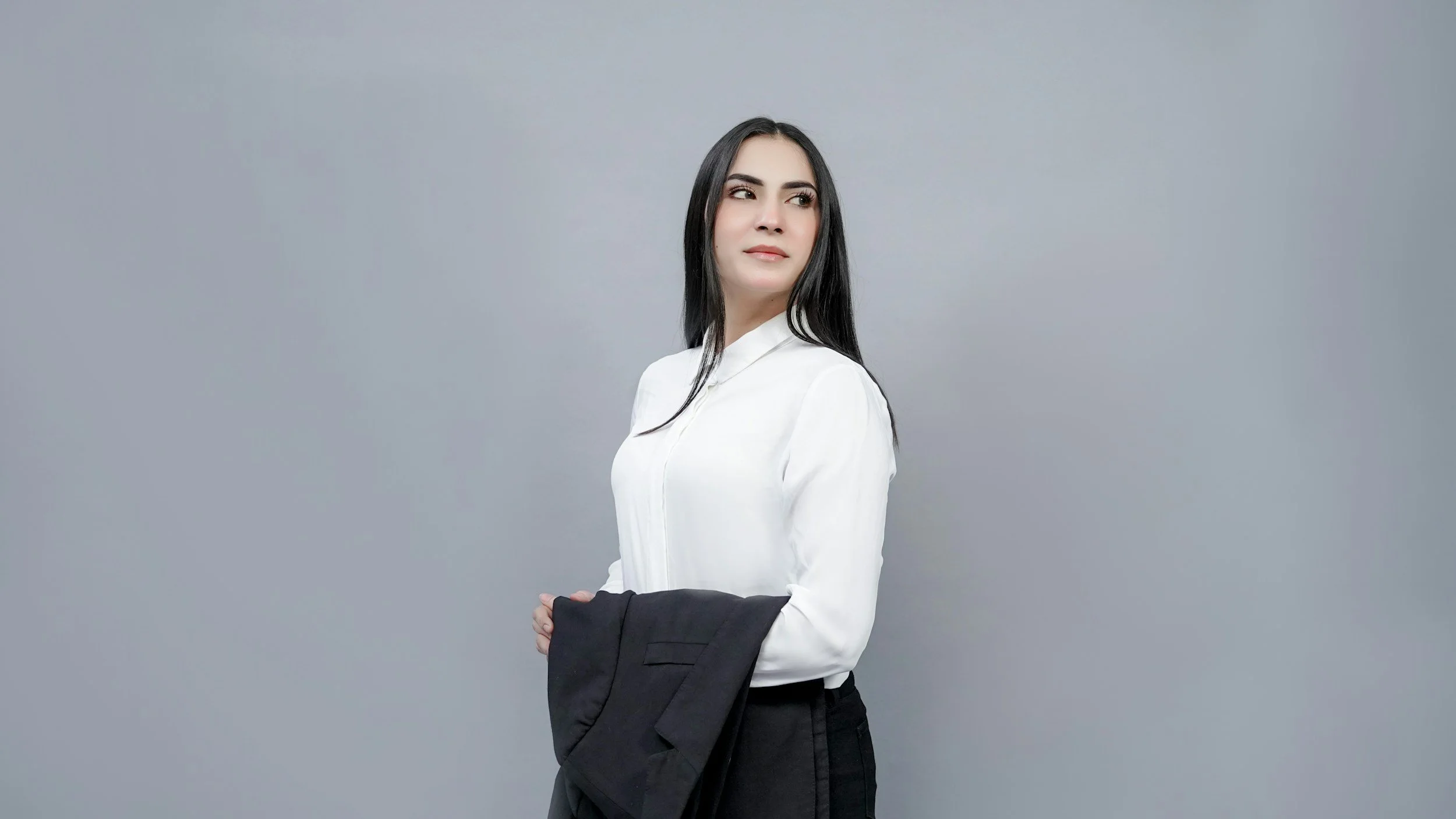 A woman with long dark hair and fair skin stands against a plain grey background, wearing a white blouse and black pants, holding a black blazer in her left hand, looking to her left with a contemplative expression.