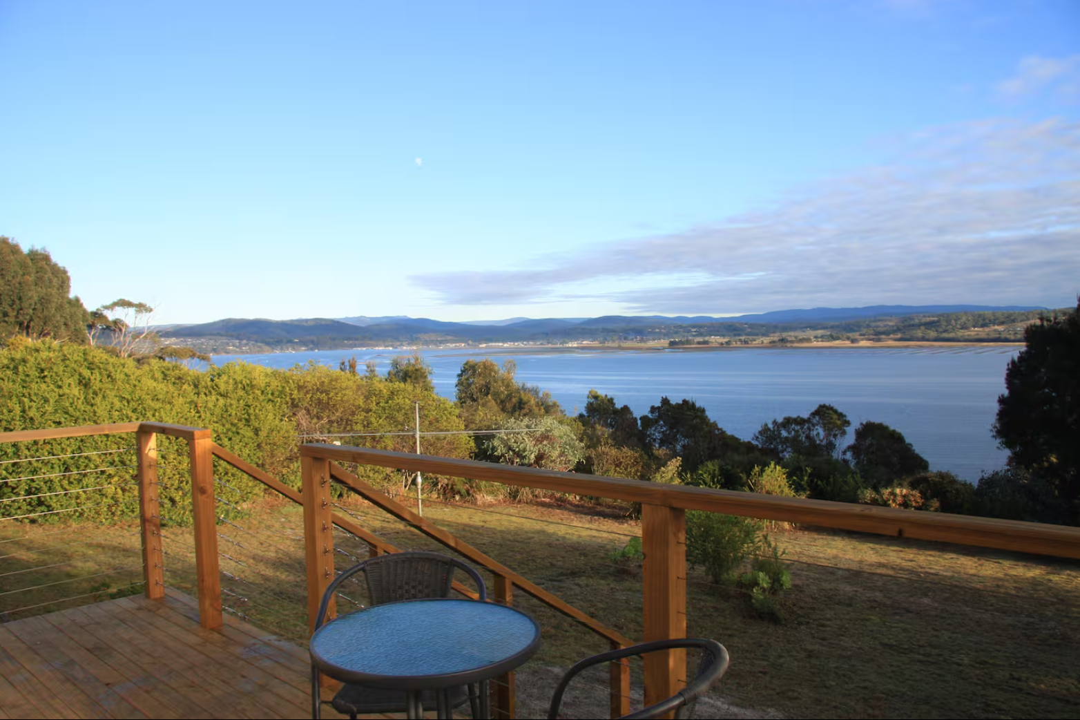 A large, two-story house with a gray shingle roof, white porch railing, and multiple windows, surrounded by green lawn and trees in autumn.