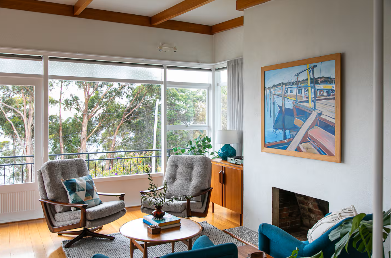 Living room with large window view of trees, light wood furniture, two gray armchairs with a pillow, a blue armchair, a wooden coffee table with books, a brick fireplace, and a framed painting of boats on the wall.