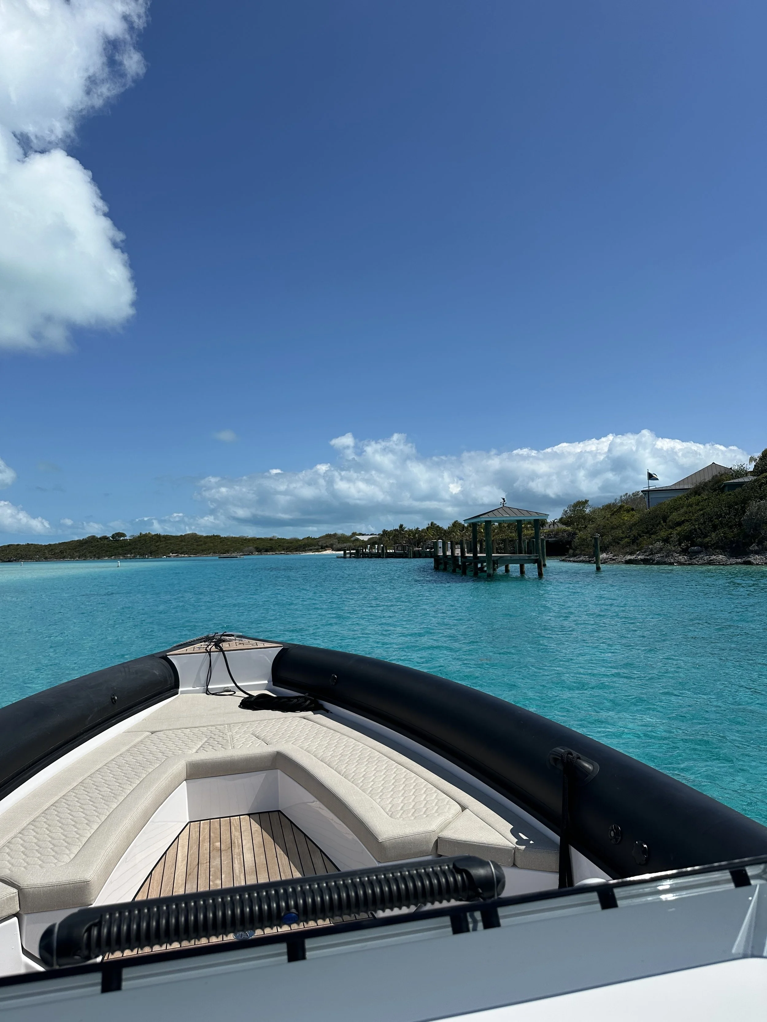 View from a boat facing a turquoise waterway with a small pier with a gazebo and some buildings on the horizon, under a blue sky with scattered clouds.