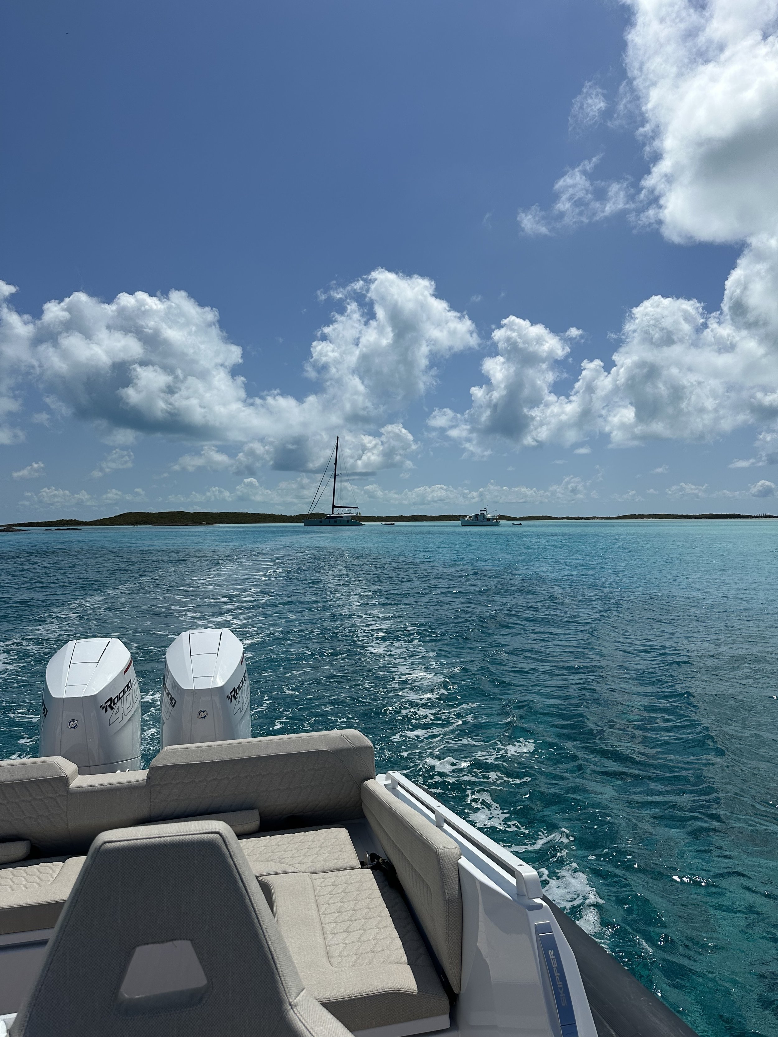 View from the back of a boat showing two white outboard engines, with a seating area in the foreground, moving through deep blue water towards a horizon with sailboats and a partly cloudy sky.