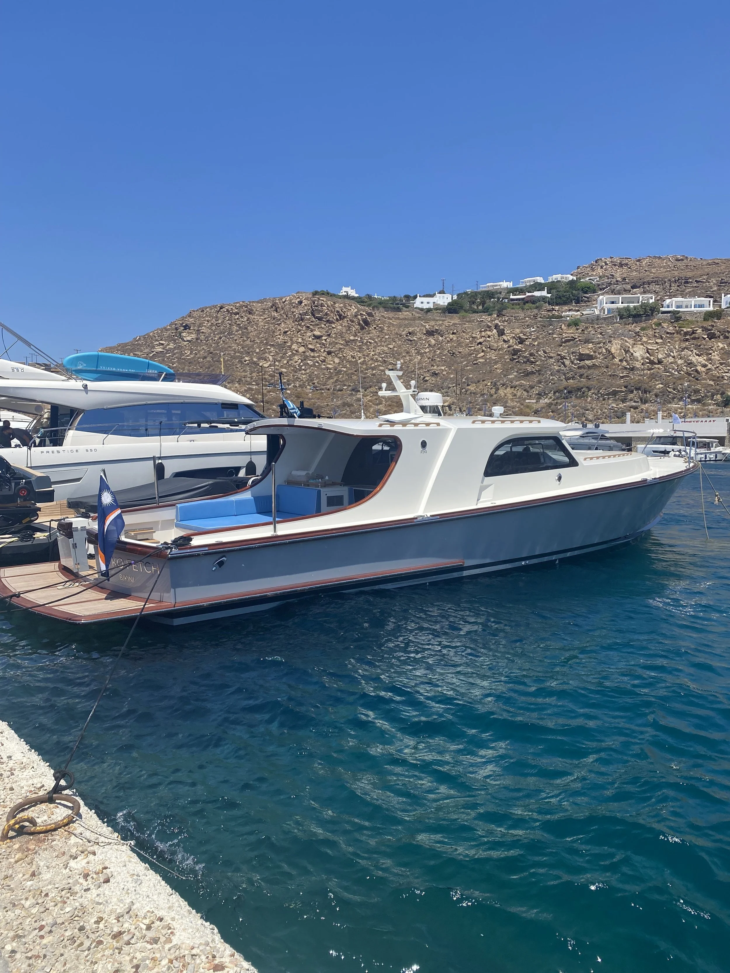 White luxury yacht docked at a marina with other boats, rocky hillside with white buildings in the background, blue sky, and clear water in the foreground.
