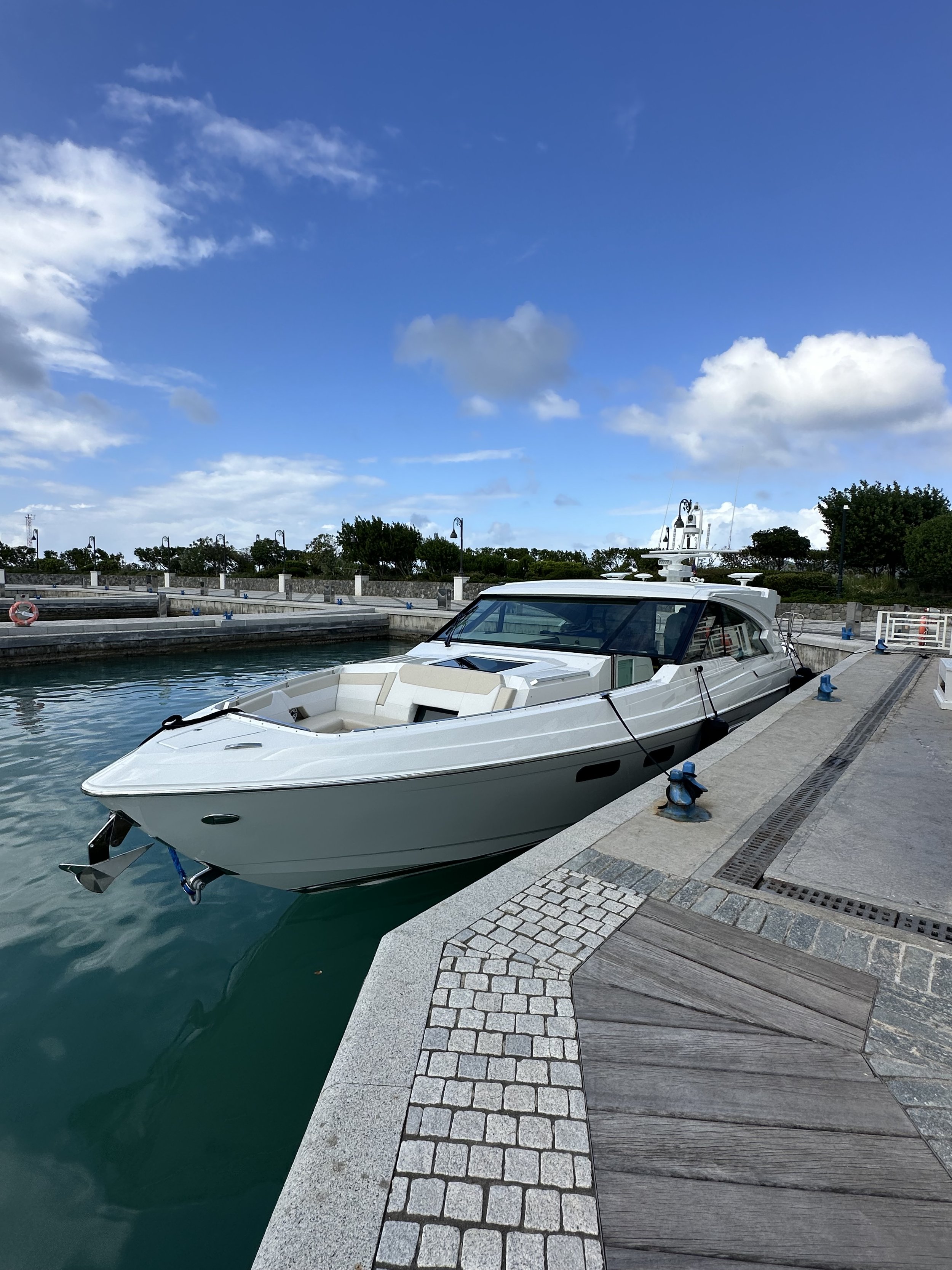 A white yacht docked at a marina with a stone and wooden pier. The sky is partly cloudy with a blue background.