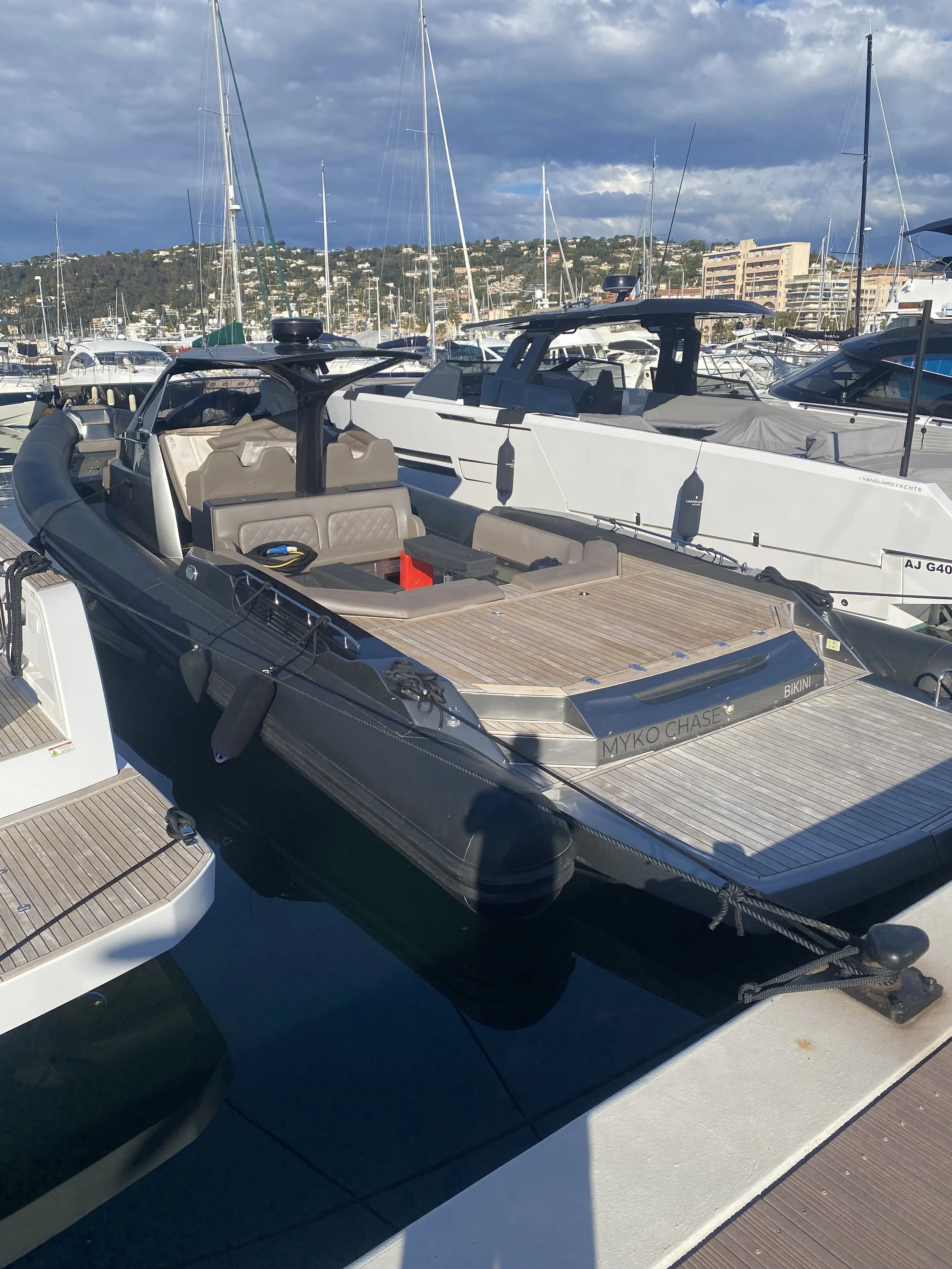 A luxurious yacht docked at a marina with other boats, hills, and buildings in the background under partly cloudy skies.