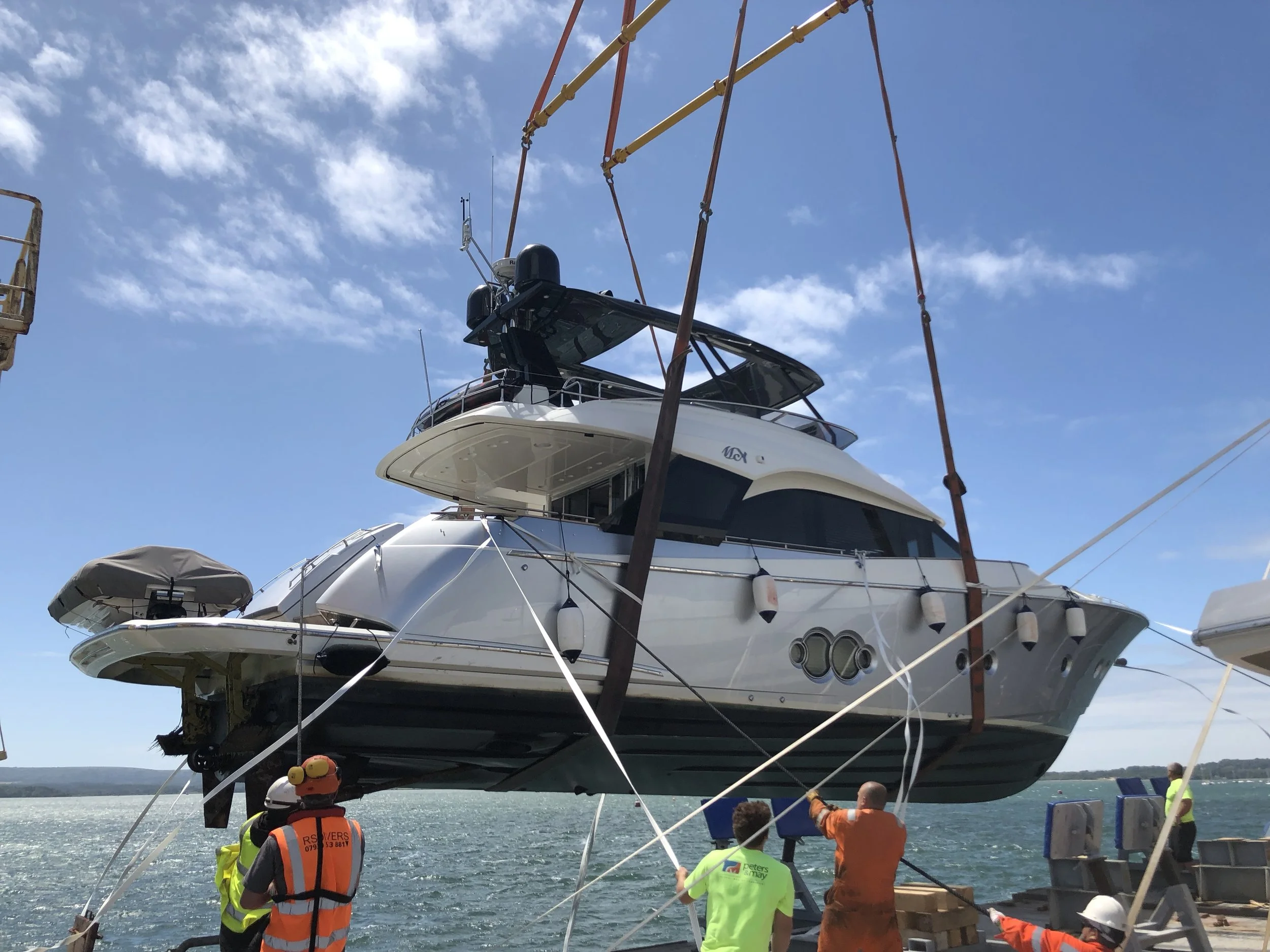 A large luxury yacht being lifted by crane onto a boat dock with workers guiding it, under a partly cloudy sky.