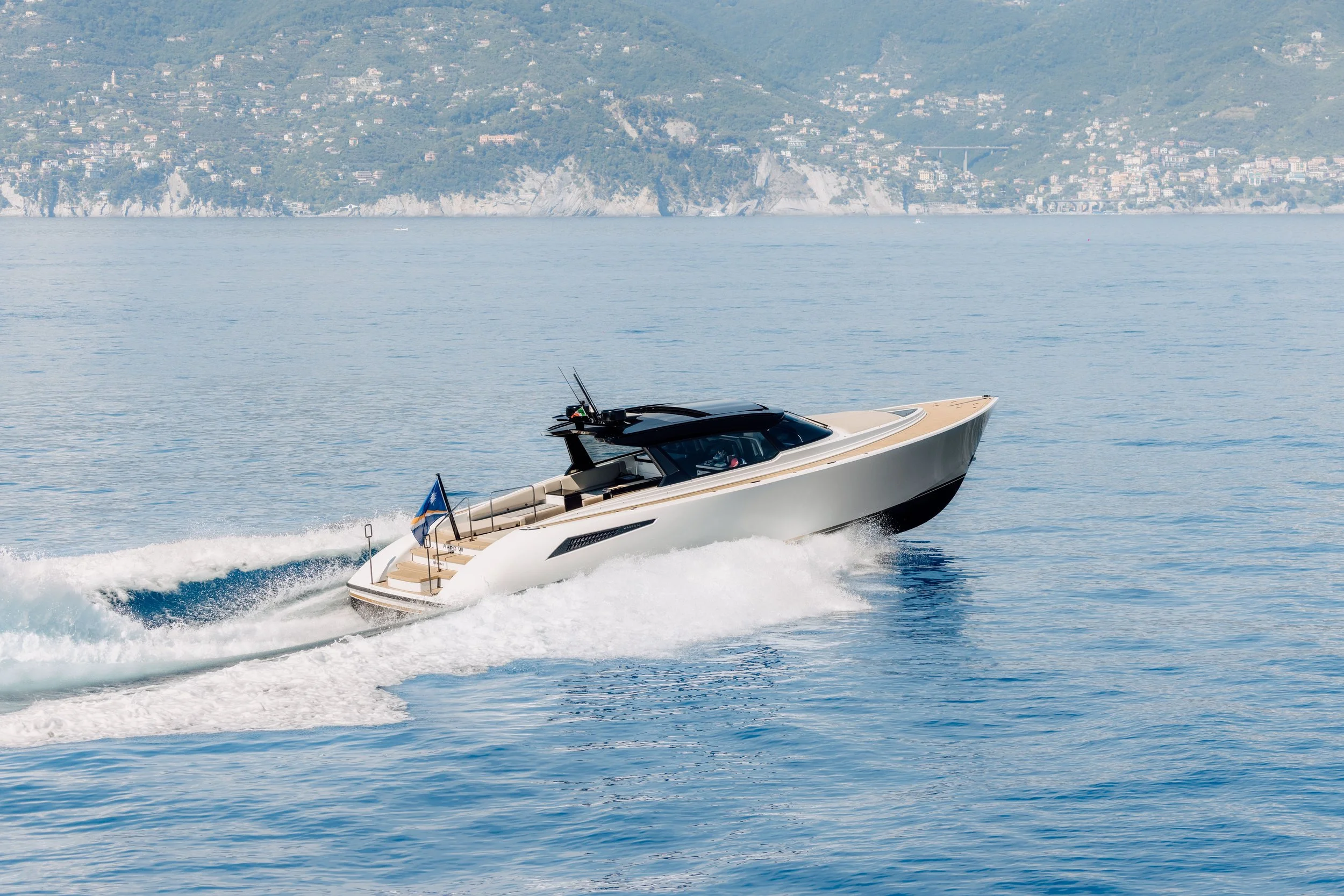 A luxury motor yacht speeding across calm blue water with a city and hillside in the background.