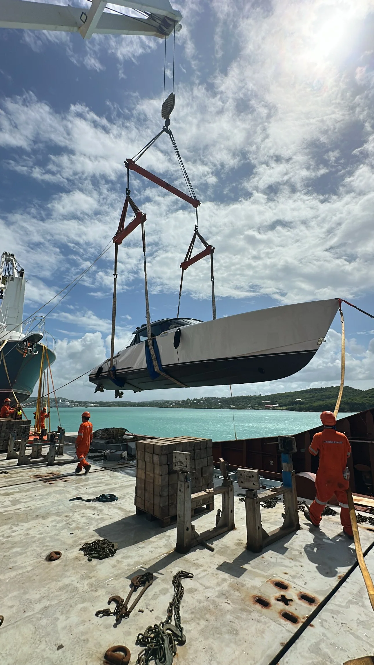 Workers in orange uniforms and helmets are lifting a boat with a crane at a dock, with blue water and a partly cloudy sky in the background.