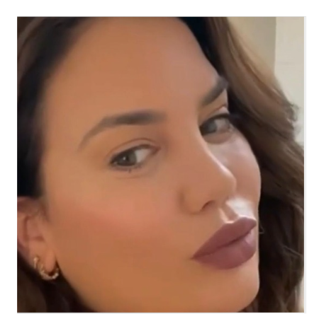 Close-up portrait of a woman with long dark wavy hair, wearing makeup with neutral lipstick, earrings, and a fur coat against a peach background. The name 'Suzanne' is displayed in an orange label at the bottom.