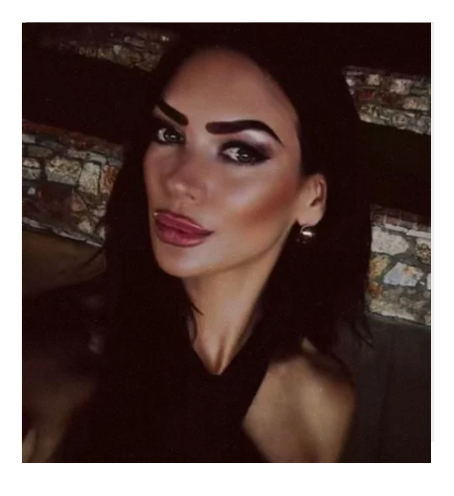 A woman with long, wavy brown hair and makeup, wearing a sleeveless black top, stands against a gradient peach and yellow background.