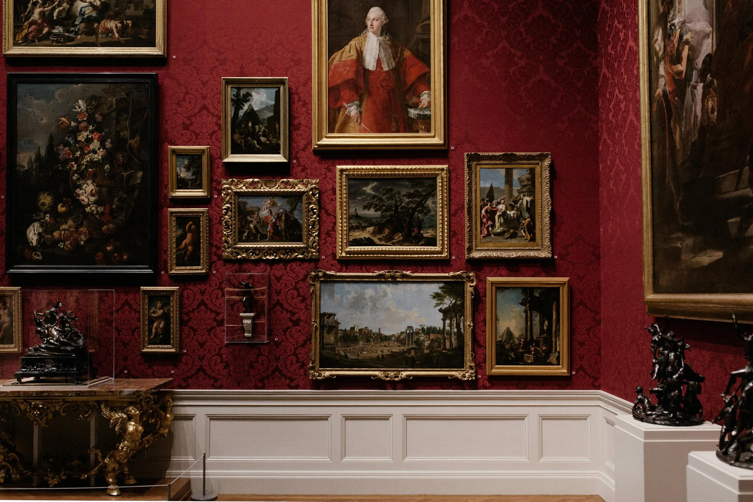 A gallery wall with multiple framed classical paintings on red patterned wallpaper, including portraits, landscapes, and still life, with sculptures on display below.
