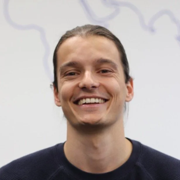 Smiling man with long brown hair pulled back, wearing a dark sweater, standing in front of a whiteboard with faint drawing lines.