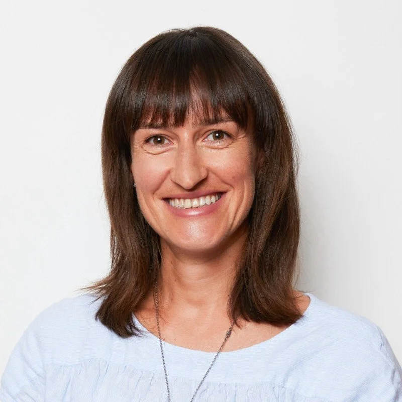 Portrait of a woman with shoulder-length brown hair and bangs, smiling, wearing a light blue top, against a plain white background.