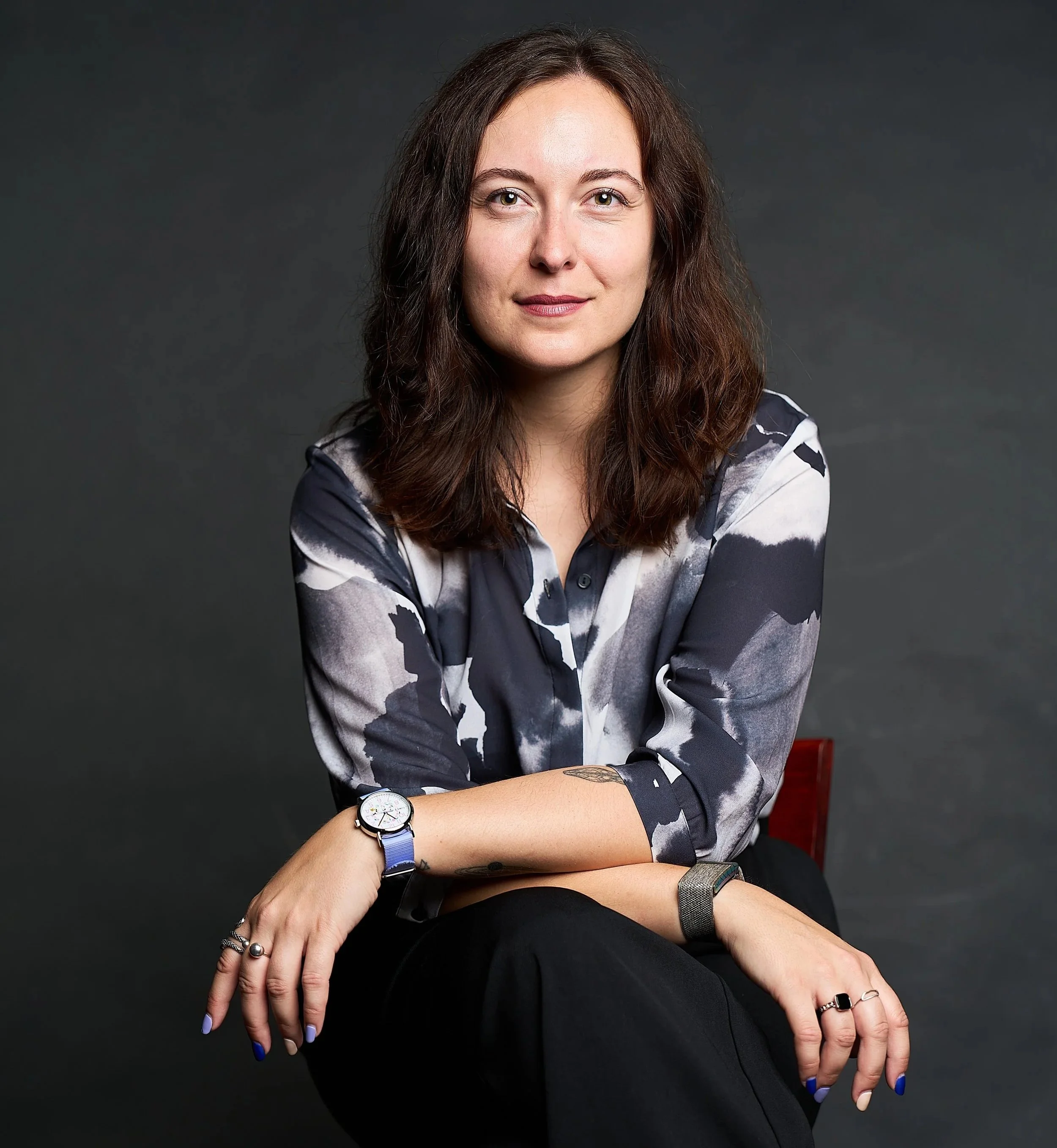 A woman with shoulder-length brown hair wearing a black, white, and gray patterned blouse, sitting against a dark gray background.
