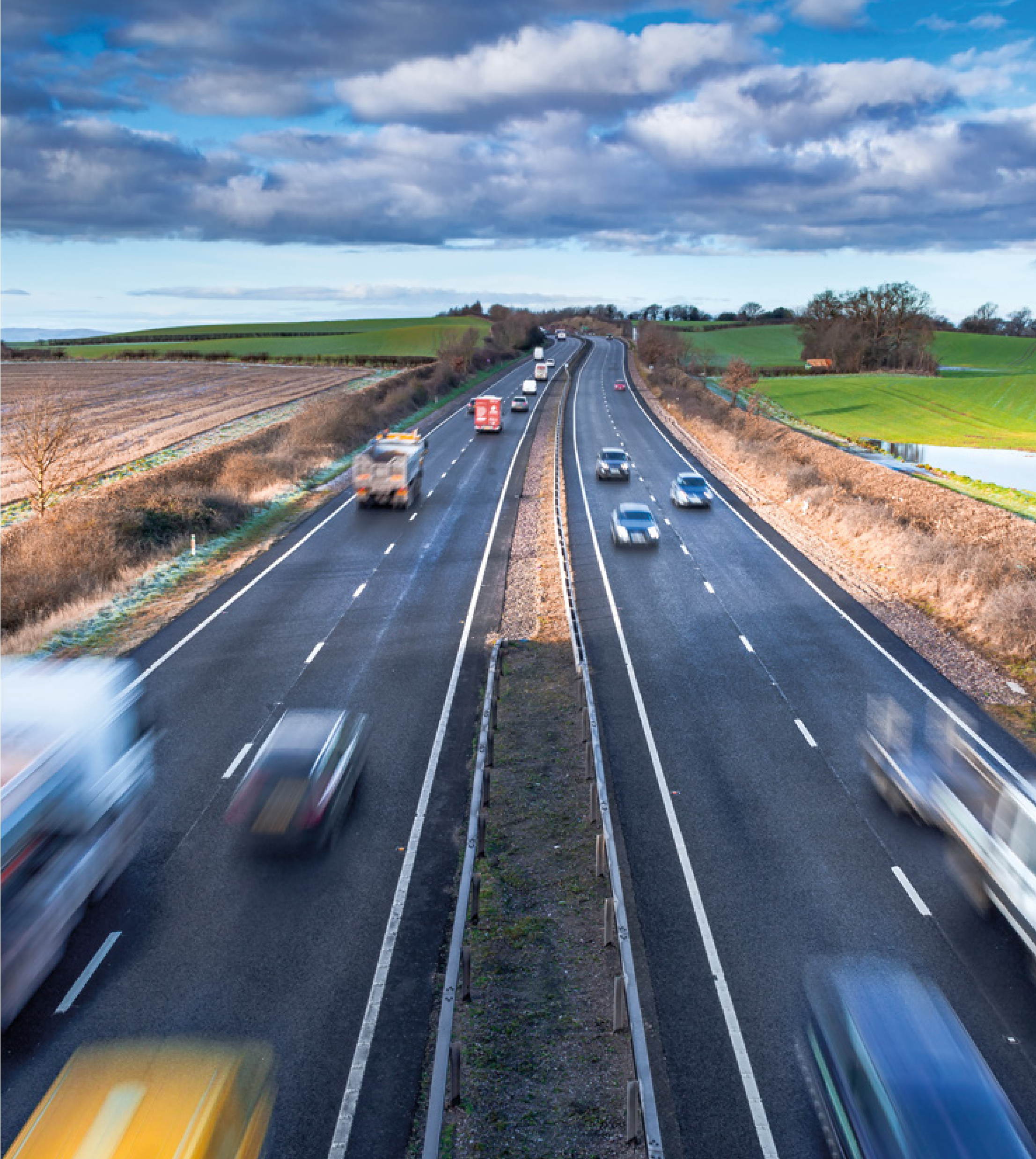 View of a highway with multiple cars and trucks traveling in both directions through a rural area with fields and trees under a cloudy sky.
