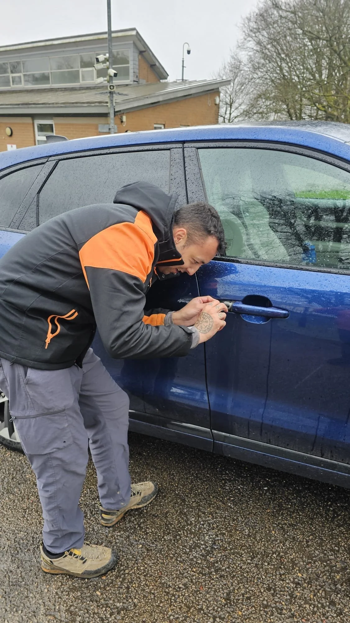 A man in a black and orange jacket leaning over a blue car, inspecting or working on the lock on the driver's side door with a tool, on a rainy day.