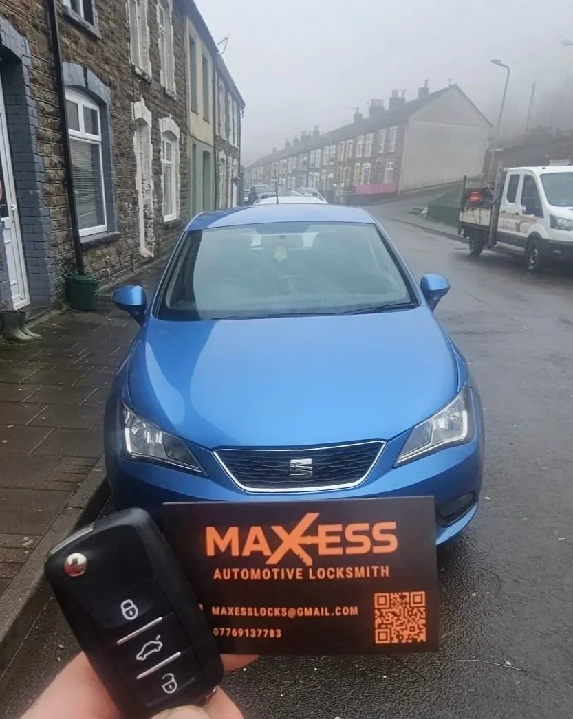 A blue car parked on a misty street with brick houses in the background. Someone is holding a car key and business card for Maxess Automotive Locksmith in front of the car.