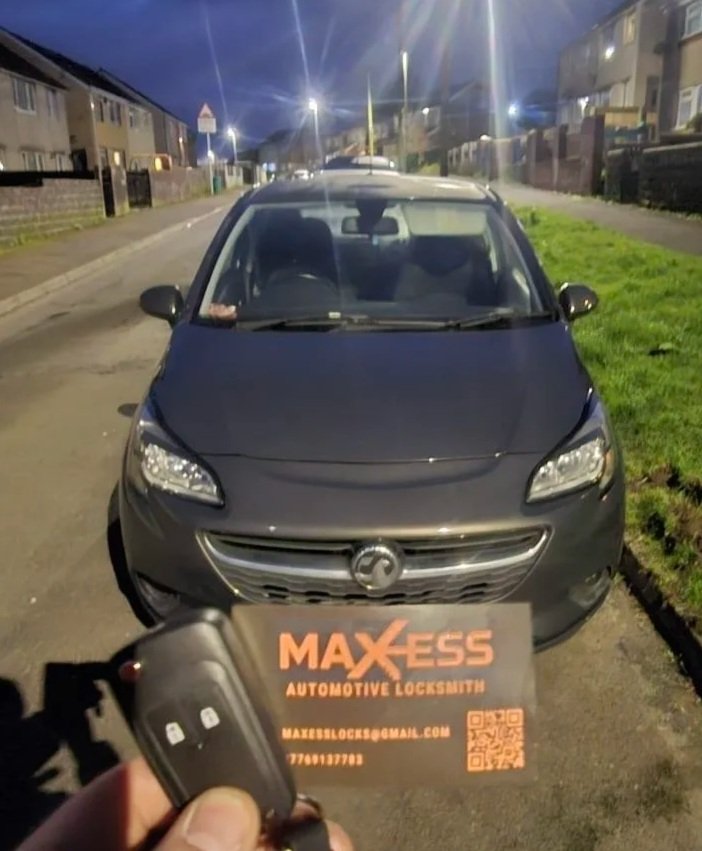 A gray car parked on the street at night with streetlights and houses in the background. The photo is taken from the front, showing the car's headlights, grille, and windshield. In the foreground, a person is holding a car key and a sign that reads '